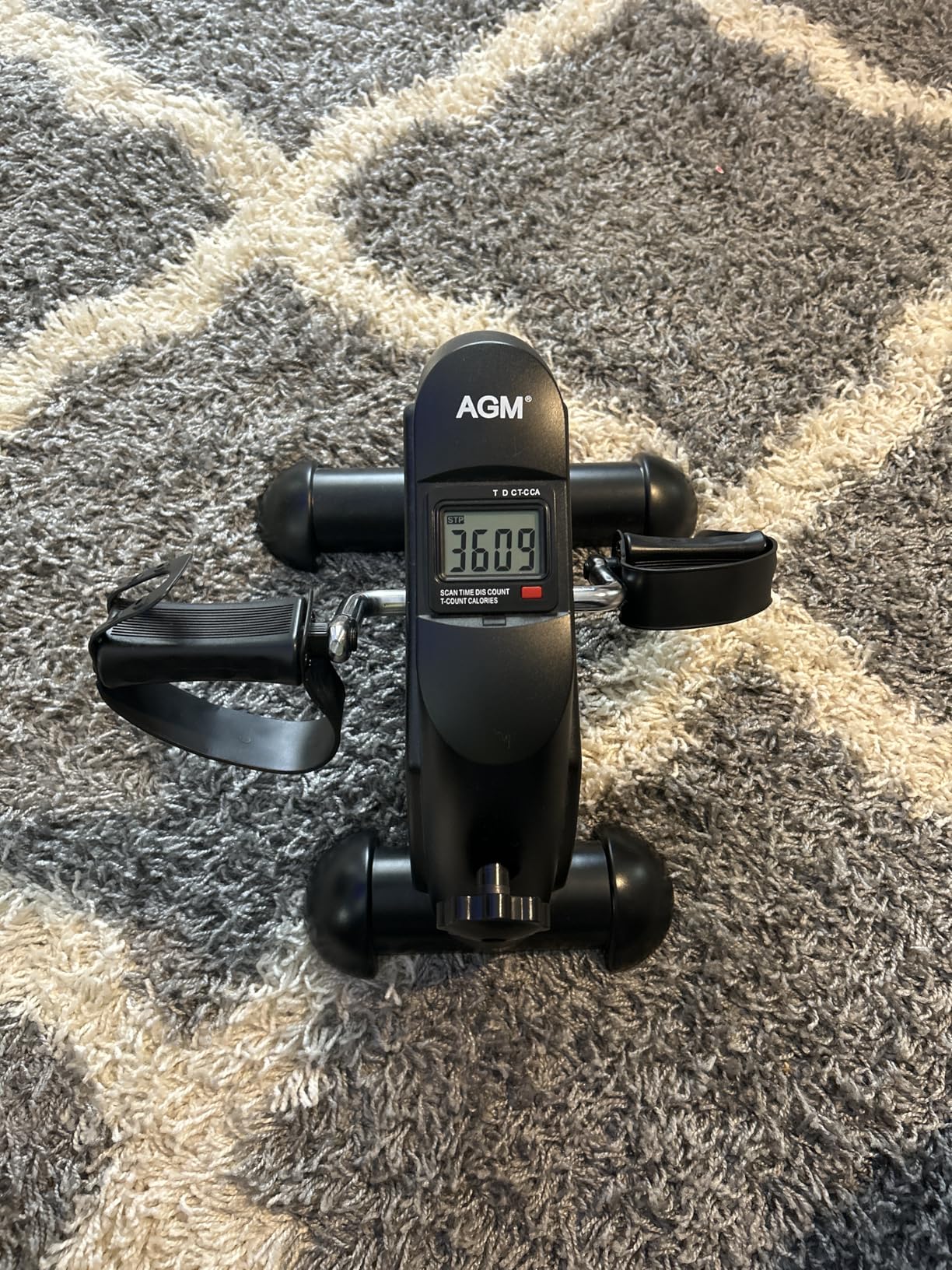 Black AGM mini exercise bike on a gray patterned rug, seen from above with the LCD monitor lit