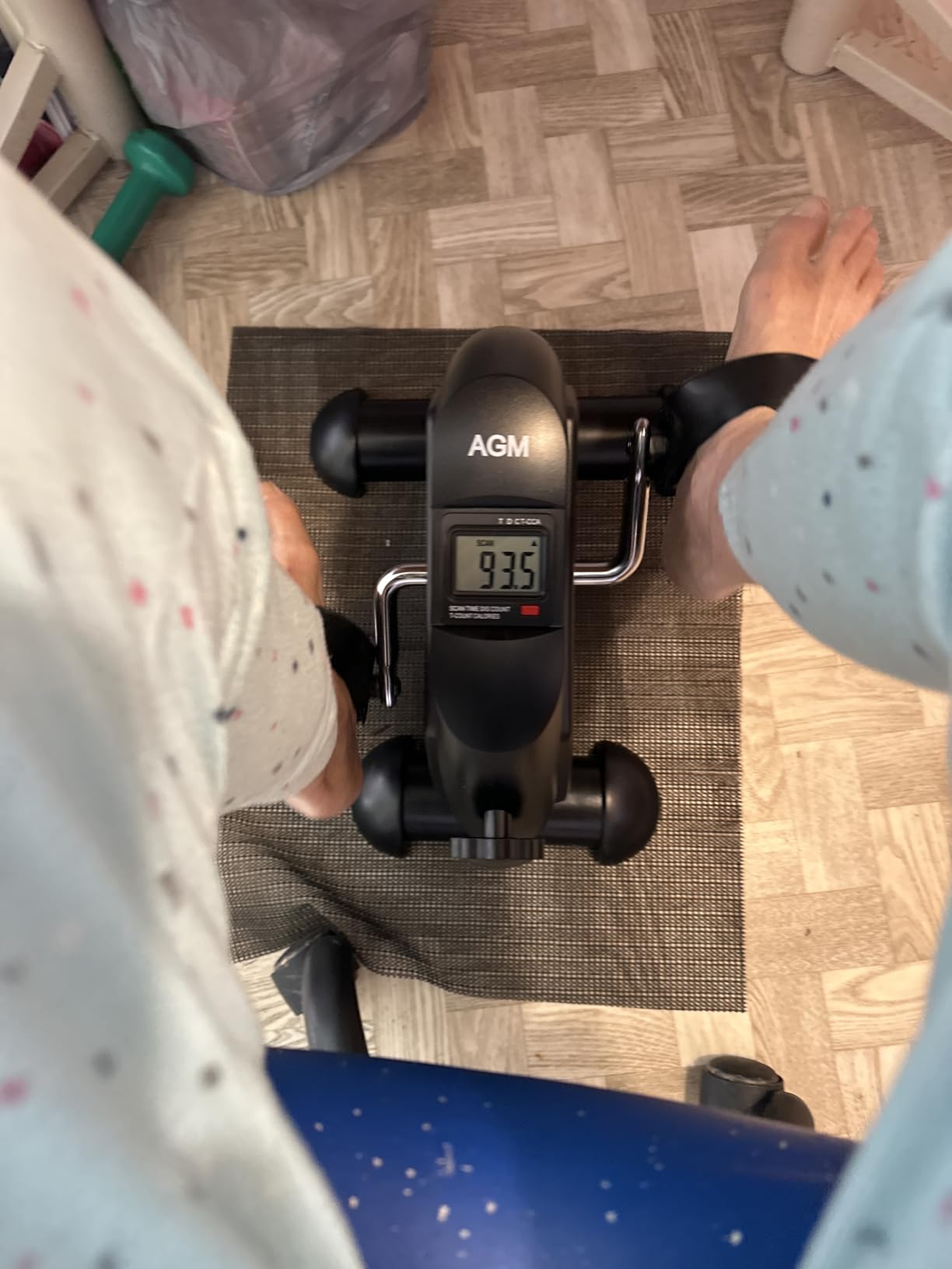 Black AGM pedal exerciser on a floor mat in front of a chair, with the buyer’s feet beside it
