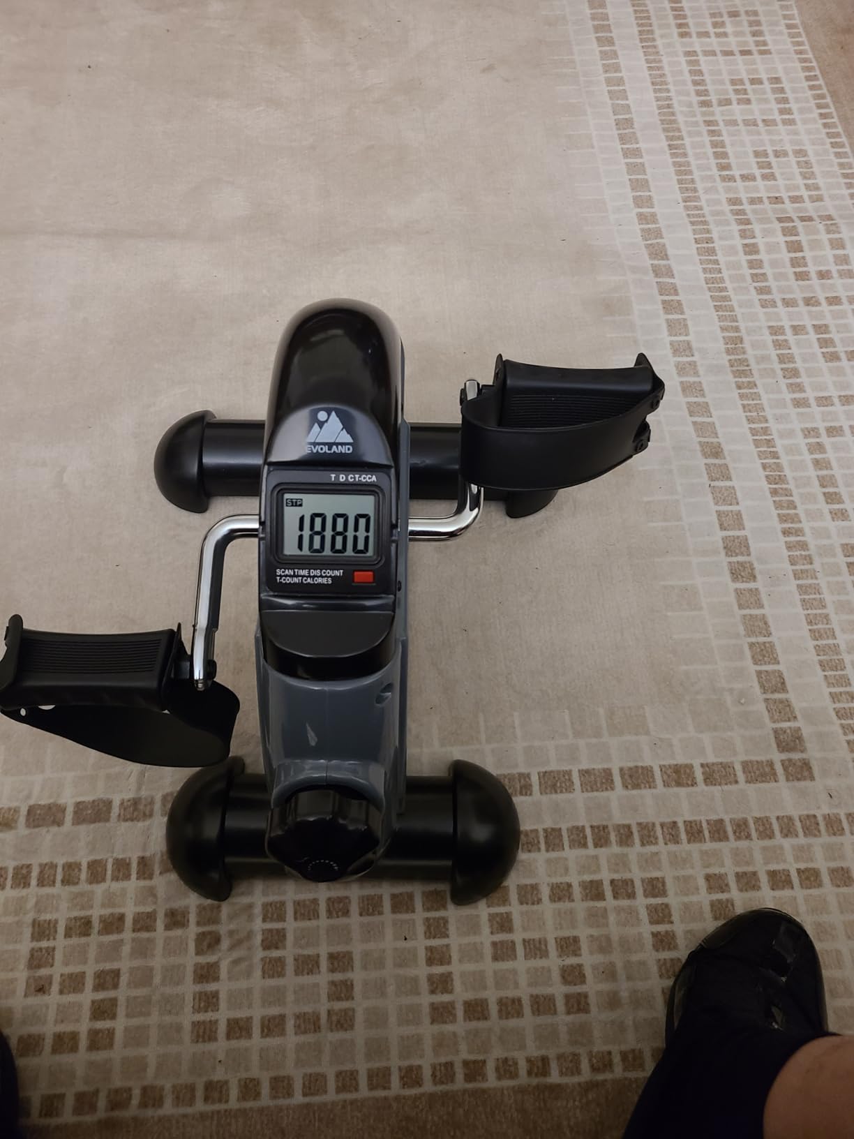 Gray and black mini pedal exerciser with strapped pedals and an LCD monitor on a beige rug