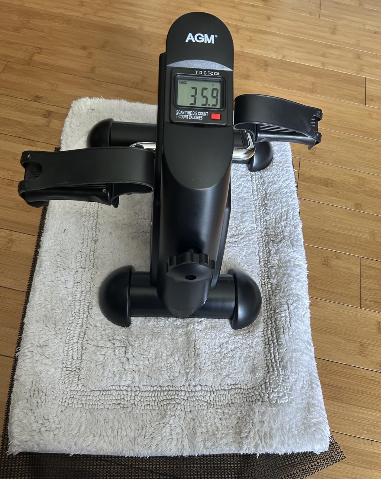 Black AGM mini exercise bike on a white mat over a wood floor, with the LCD monitor showing a reading