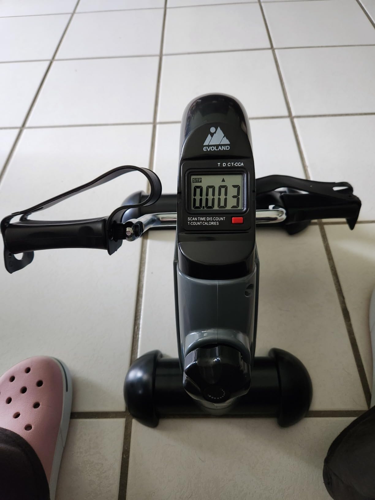 Black mini pedal exerciser with foot straps on a white tile floor