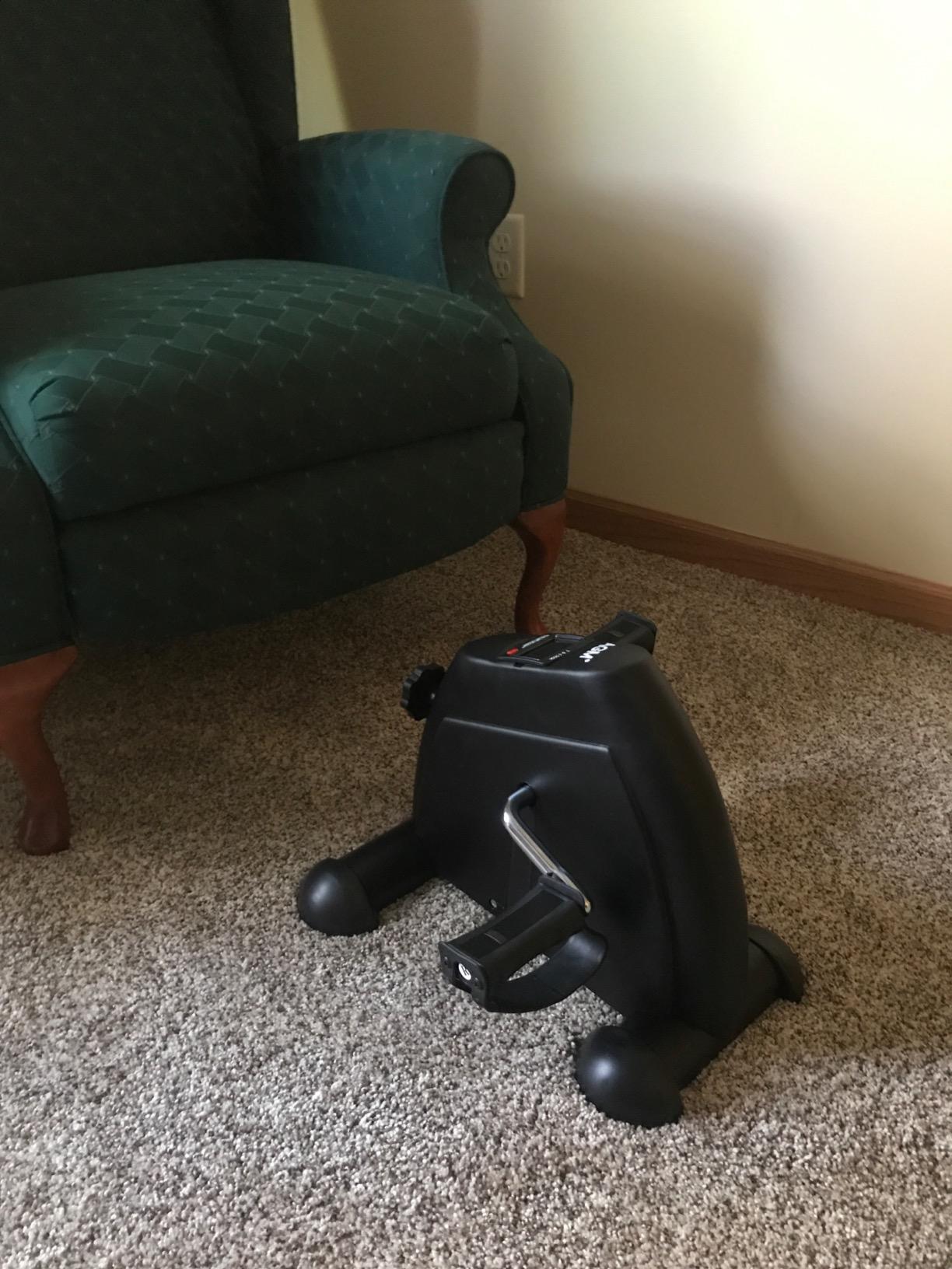 Black mini exercise bike on carpet in front of a green armchair